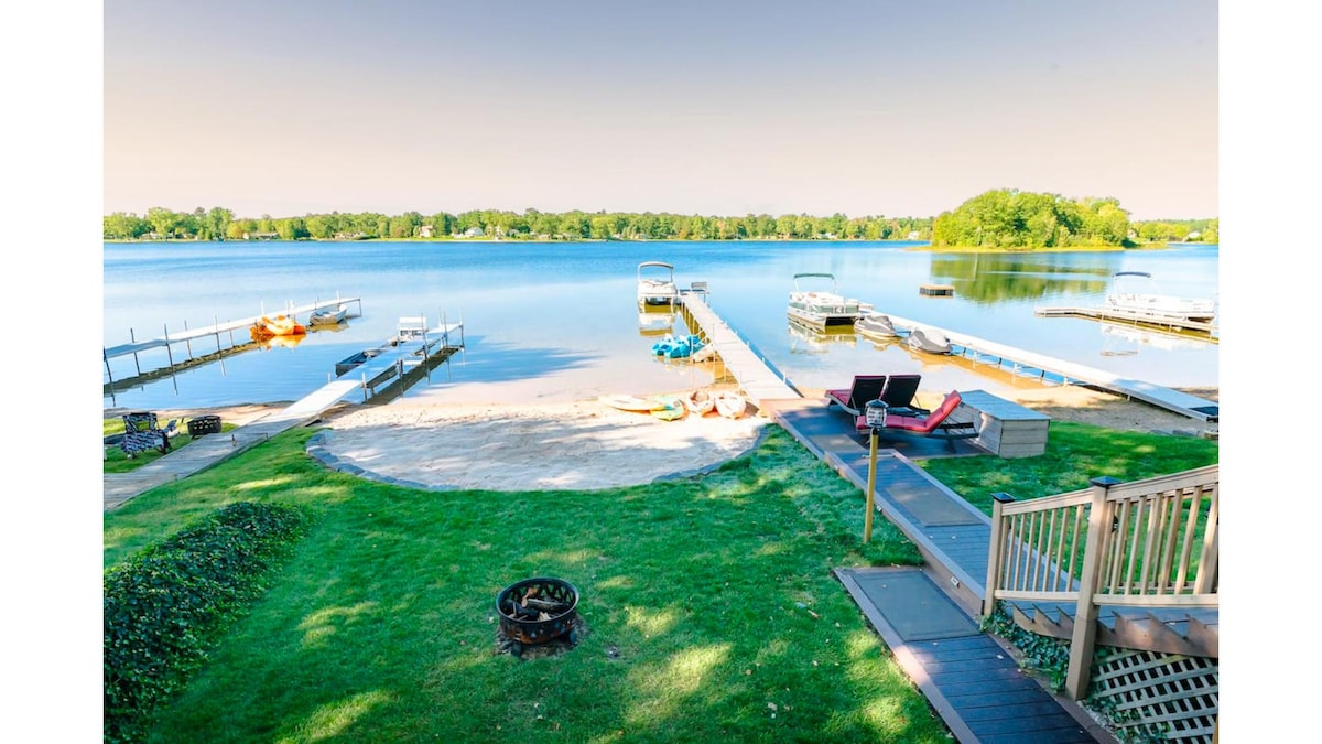 A serene lakefront scene captures the calm waters reflecting light. A private sandy beach is visible, accompanied by multiple docks and boats. Lounge chairs are arranged on the grass, inviting relaxation, while a fire pit offers a cozy gathering spot.