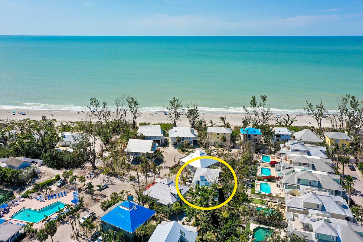 An aerial view showcases the stilted home nestled within the Sunset Captiva community. The property is surrounded by greenery, with the beach and calm ocean visible in the background. Nearby amenities like the pool and tennis courts are also highlighted.
