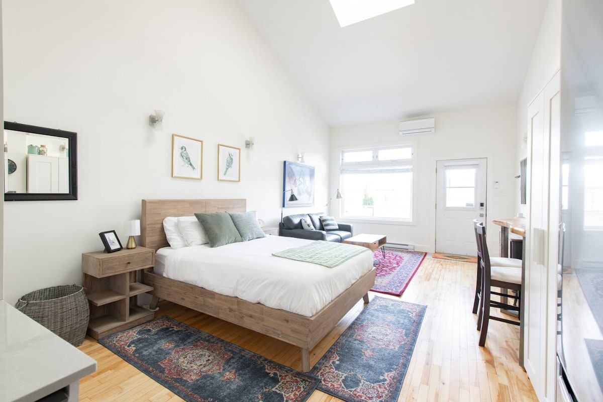 A bright studio room features high ceilings and large windows allowing for abundant natural light. A spacious bed is positioned beside a comfortable couch, with a small table and two chairs set nearby. Decorative artwork adorns the walls, complementing the hardwood flooring.