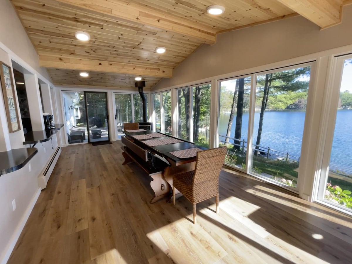 A sunlit room features large glass windows that offer views of Little Wolf Lake. A wooden table occupies the center, surrounded by woven chairs. Pine ceilings and hardwood flooring add warmth to the space. A fireplace stands at the far end, enhancing the room's inviting atmosphere.