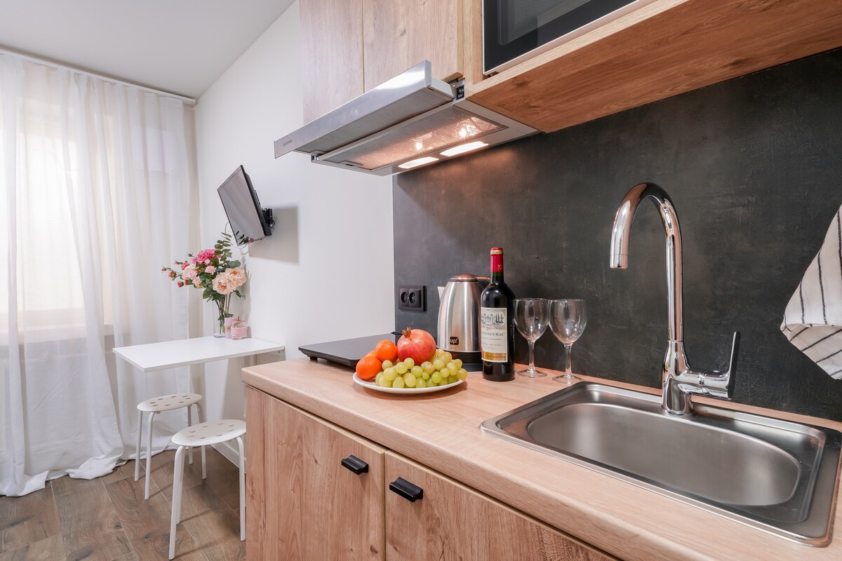 A modern kitchen area is shown with wooden cabinetry and a stainless-steel sink. A platter of fresh fruits sits next to a bottle of wine and glasses. A small table with two white chairs is visible, alongside a wall-mounted television and a bouquet of flowers.