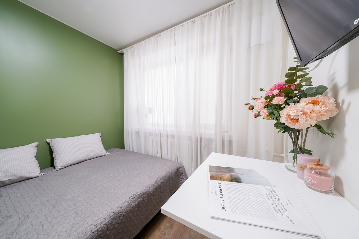 A bright sleeping area features a spacious bed covered with a gray quilt. Soft light filters through sheer white curtains, illuminating the room. A small table holds a decorative flower arrangement and a magazine, complemented by a mounted television on the wall.