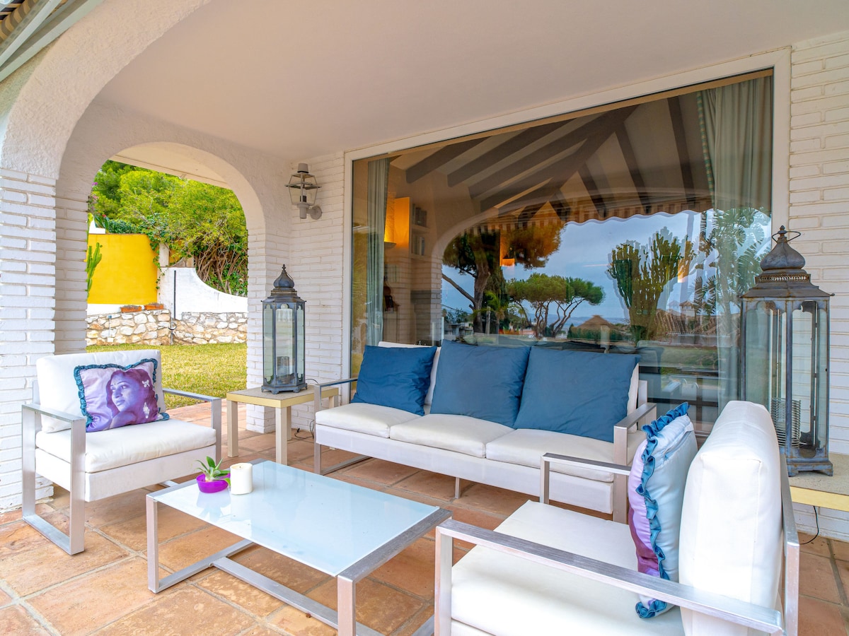 A covered outdoor seating area features a white sofa and two armchairs adorned with blue cushions. A glass-top table sits in front, complemented by decorative lanterns on either side. The space is framed by arches and offers a view of the surrounding garden.