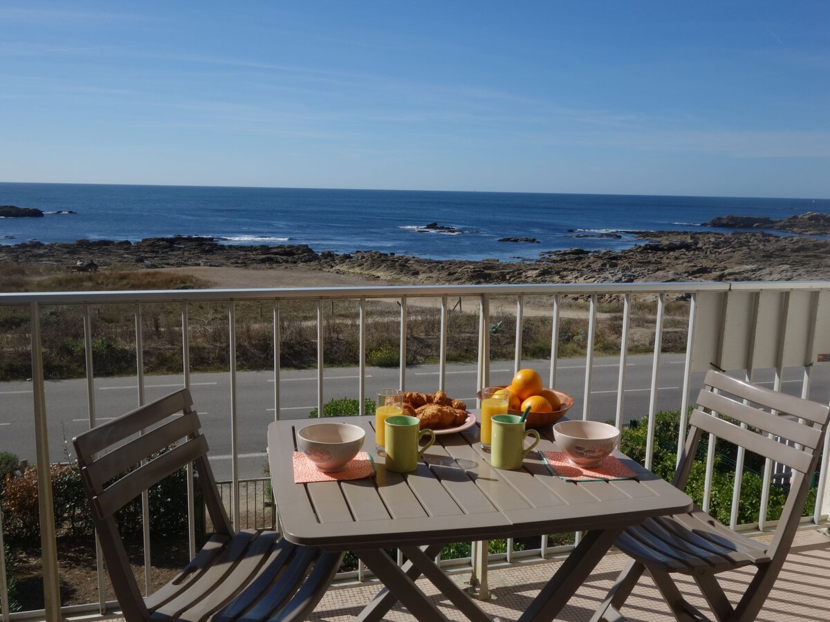 A balcony table set for breakfast is displayed, featuring two green mugs, pastries, and a bowl of fruit. The ocean is visible in the background with rocky formations, creating a scenic coastal view. Sunny skies and gentle waves enhance the tranquil ambiance.