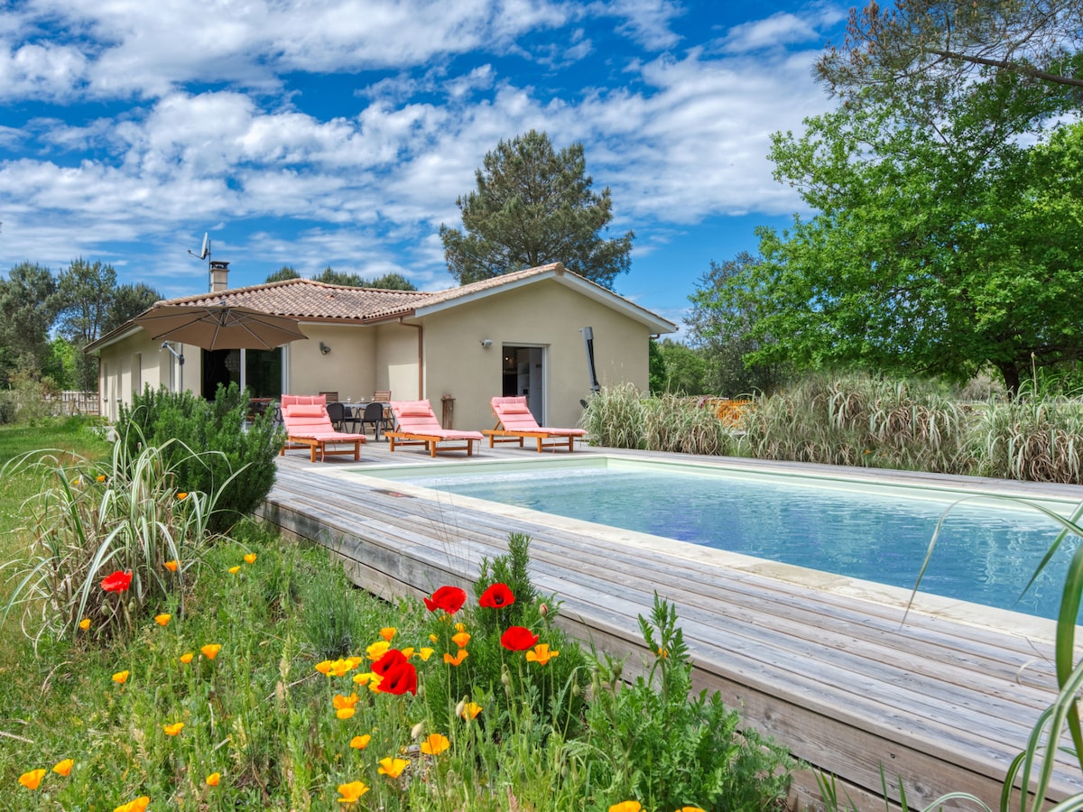 A spacious villa features a wooden deck surrounding a swimming pool, accentuated by colorful flowers in the foreground. Sun loungers are positioned nearby under an umbrella, with greenery and trees providing a natural backdrop. A clear sky with scattered clouds enhances the setting.