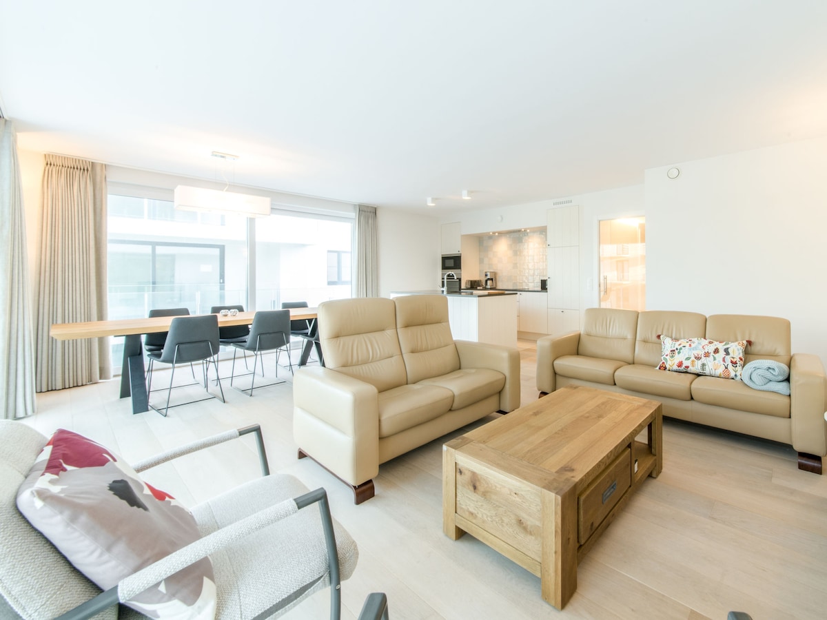 The living and dining area is characterized by neutral tones and modern furniture. Comfortable leather sofas face a wooden coffee table, while a large dining table accommodates multiple chairs. Natural light floods the space through expansive windows leading to the balcony.