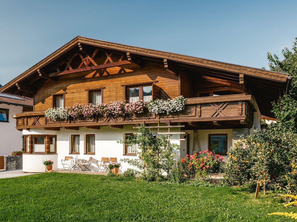 A two-story wooden chalet is surrounded by vibrant greenery and flower beds. A spacious balcony adorned with flowers extends from the upper level. A grassy area is visible in front, with a seating arrangement featuring a table and chairs. The exterior reflects traditional alpine architecture.