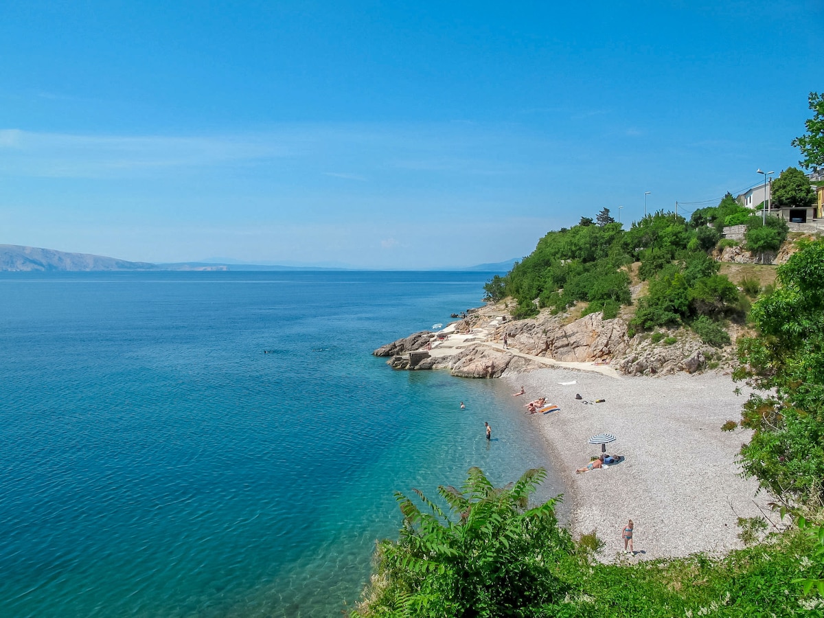 A serene pebble beach panorama features a clear blue sea gently lapping against the shore. Sunbathers and beach umbrellas are positioned along the shoreline, surrounded by lush greenery and rocky formations. The sky is bright and blue, contributing to a tranquil coastal atmosphere.