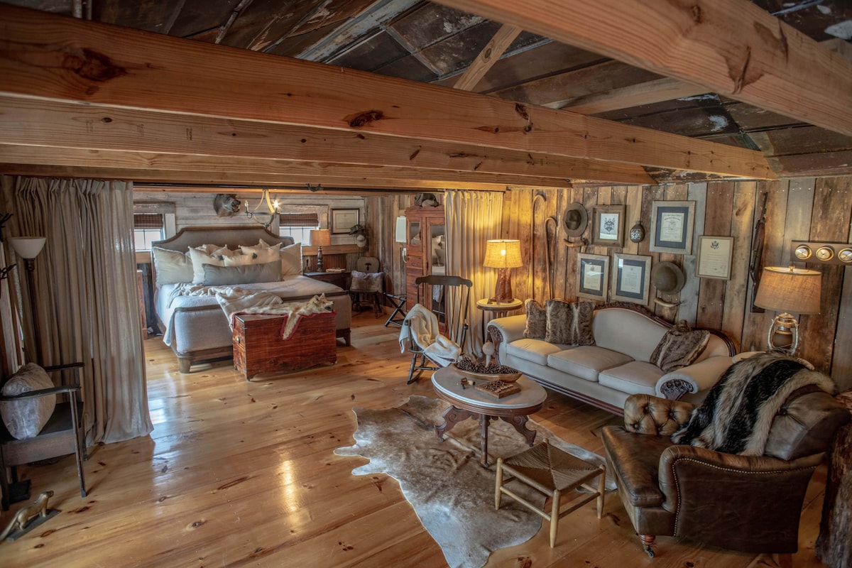 An inviting interior showcases rustic wood beams and plank flooring. A spacious bed is positioned against the wall, flanked by bedside tables. A comfortable seating area includes a sofa and an armchair, with decorative items adding character.