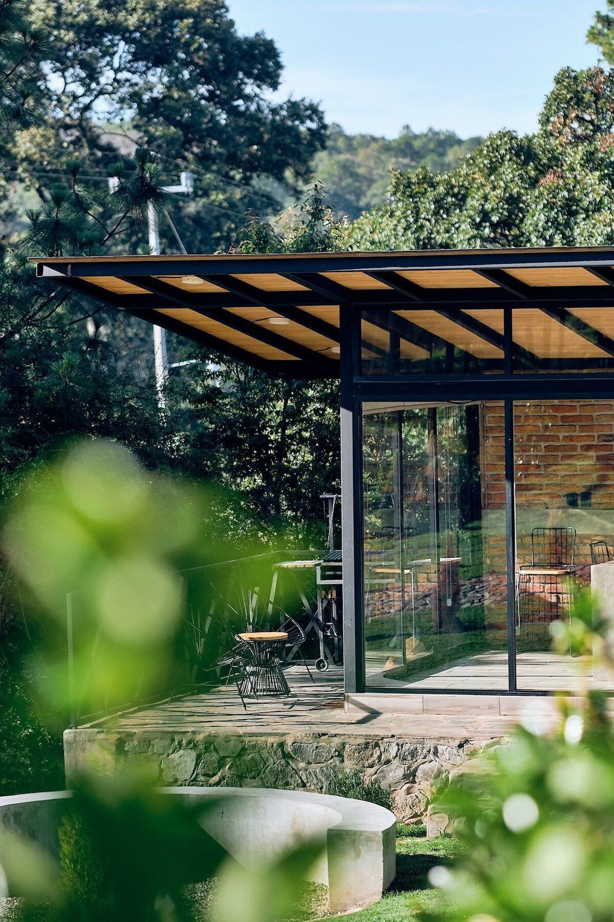 A modern cabin exterior is visible, featuring large glass panels and a wooden overhang. Surrounding greenery creates a natural frame, while a small seating area with a chair is positioned on the outdoor terrace, inviting relaxation amidst the landscape.