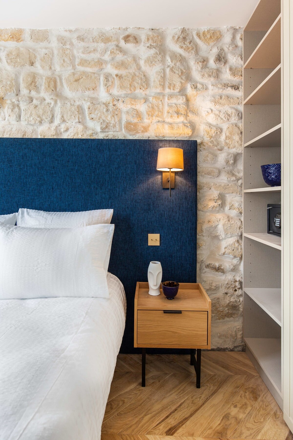 A cozy sleeping area is presented with a spacious bed featuring crisp white linens. An accent wall of textured blue fabric contrasts with the natural stone backdrop. A bedside table holds a small decorative item and a lamp, while shelving extends against the adjacent wall.