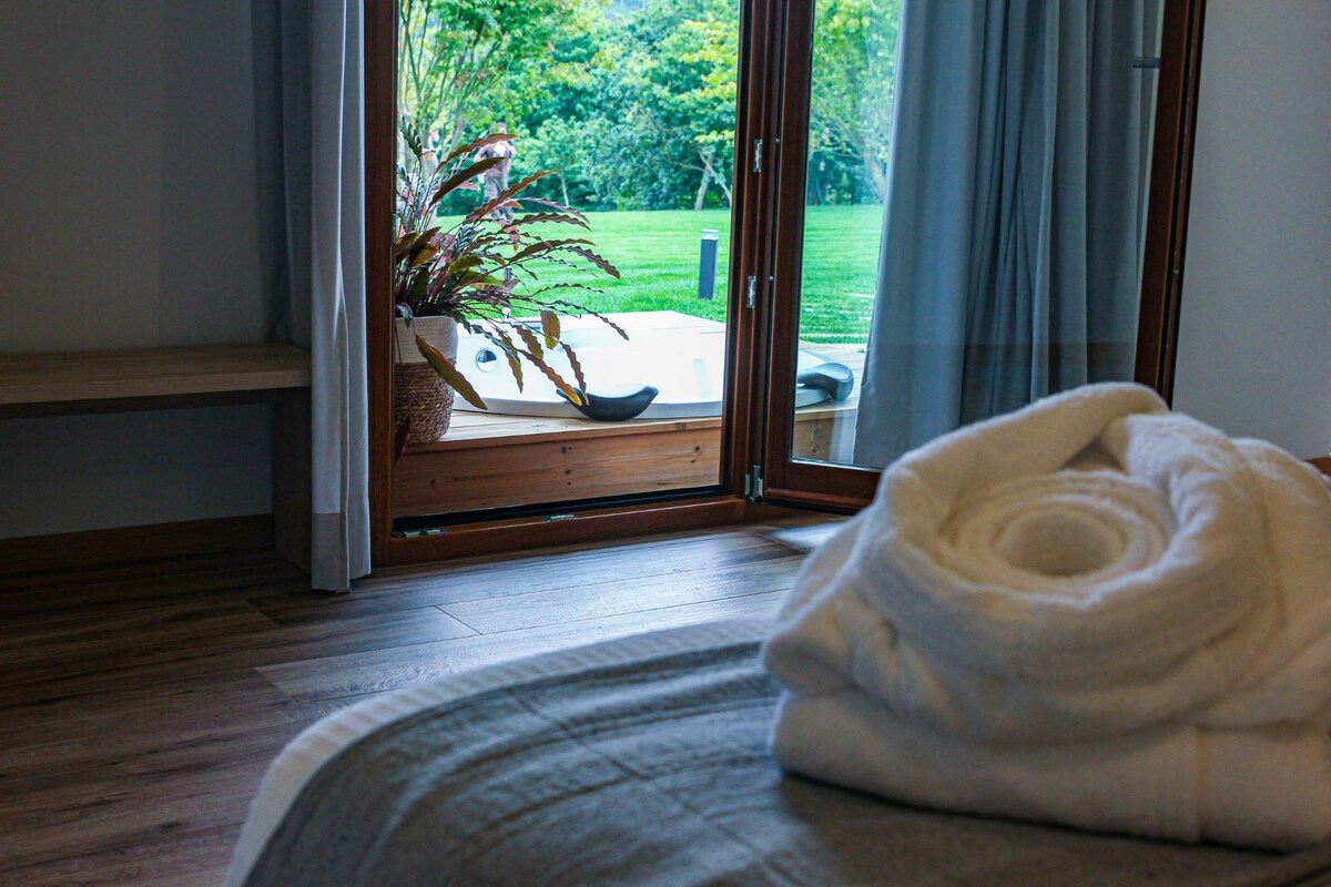 A cozy sleeping area features wooden flooring and a spacious bed with a neatly arranged towel. Glass doors open to reveal a private garden with lush greenery, and a hot tub is visible on the terrace just outside.