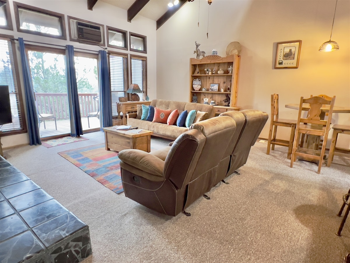 A cozy living area features a sectional sofa adorned with colorful cushions, complemented by a rustic coffee table. Natural light filters through multiple windows, enhancing the open feel. A wooden shelving unit displays various decorative items, contributing to the inviting ambiance of the space.