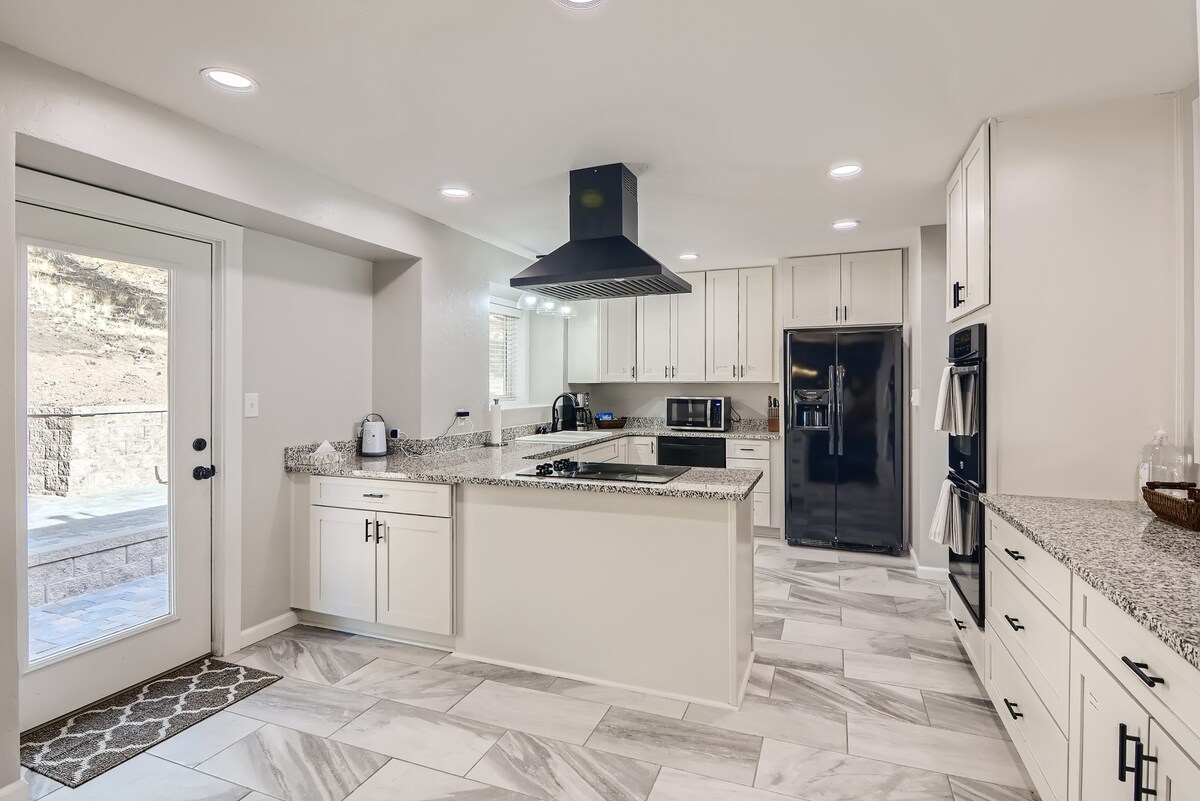 A modern kitchen is displayed with sleek cabinetry and a large island featuring a granite countertop. Stainless steel appliances include a refrigerator and double ovens. Natural light enters through a door leading to the outdoors, complemented by neutral-toned tile flooring.