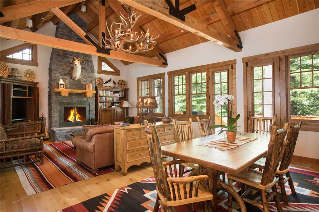 A warm living area features a stone fireplace framed by wooden beams. Rustic furniture includes a large dining table surrounded by several chairs. Natural light fills the space through multiple windows that provide views of the surrounding trees.