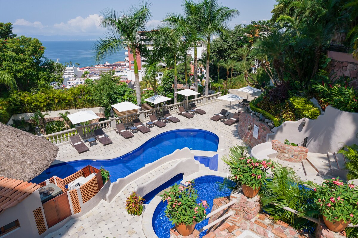 A multi-level pool area is surrounded by lush greenery and palm trees, featuring a large, inviting swimming pool with adjacent loungers. Smaller pools are integrated into the landscape, offering a serene atmosphere with views overlooking the ocean and the nearby city.