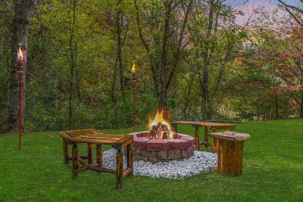 A rustic fire pit is surrounded by log seating and wooden benches, set on a bed of decorative stones. The lush greenery of trees provides a natural backdrop, and flickering flames rise from the center, creating an inviting outdoor gathering space.