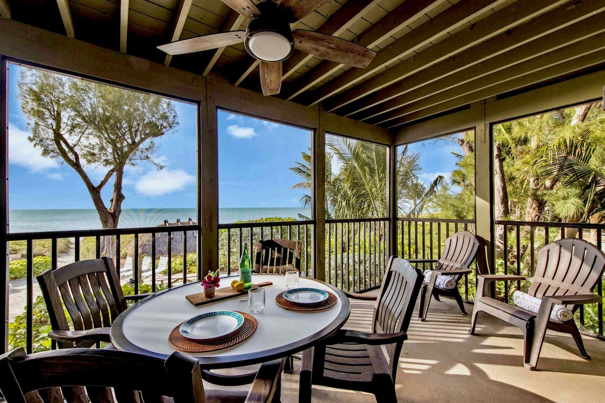 A screened porch is featured, offering an outdoor dining table set for four. Comfortable wooden chairs surround the table, with the sea and palm trees visible through large windows. Soft sunlight streams in, creating a relaxed atmosphere for enjoying meals or activities.