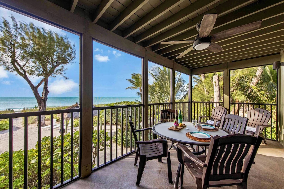 An outdoor seating area is featured on a screened porch, with a circular table surrounded by four wooden chairs. Lush greenery frames the view of the sandy beach and ocean extending towards the horizon, while a ceiling fan adds a touch of comfort.