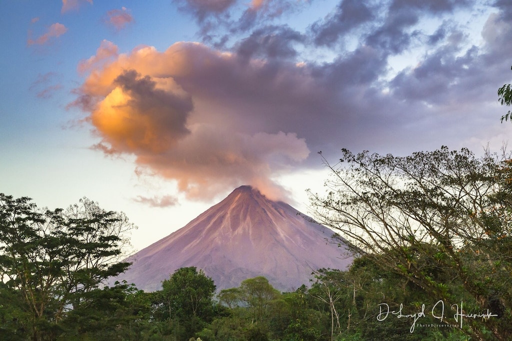 A breathtaking view of Arenal Volcano is framed by lush greenery. Soft clouds hover above the mountain, reflecting warm hues of the setting sun. The foreground features dense foliage, enhancing the vibrant natural landscape of the region.