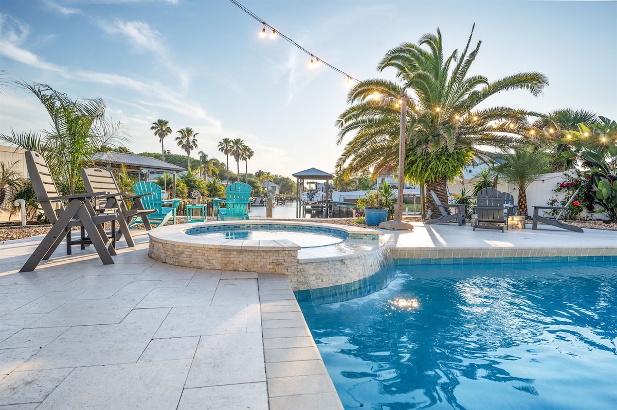 The outdoor area features a heated pool with a built-in hot tub, surrounded by a stone deck. Colorful lounge chairs and palm trees create a tropical ambiance. Soft lighting is strung overhead, enhancing the tranquil setting by the canal at sunset.