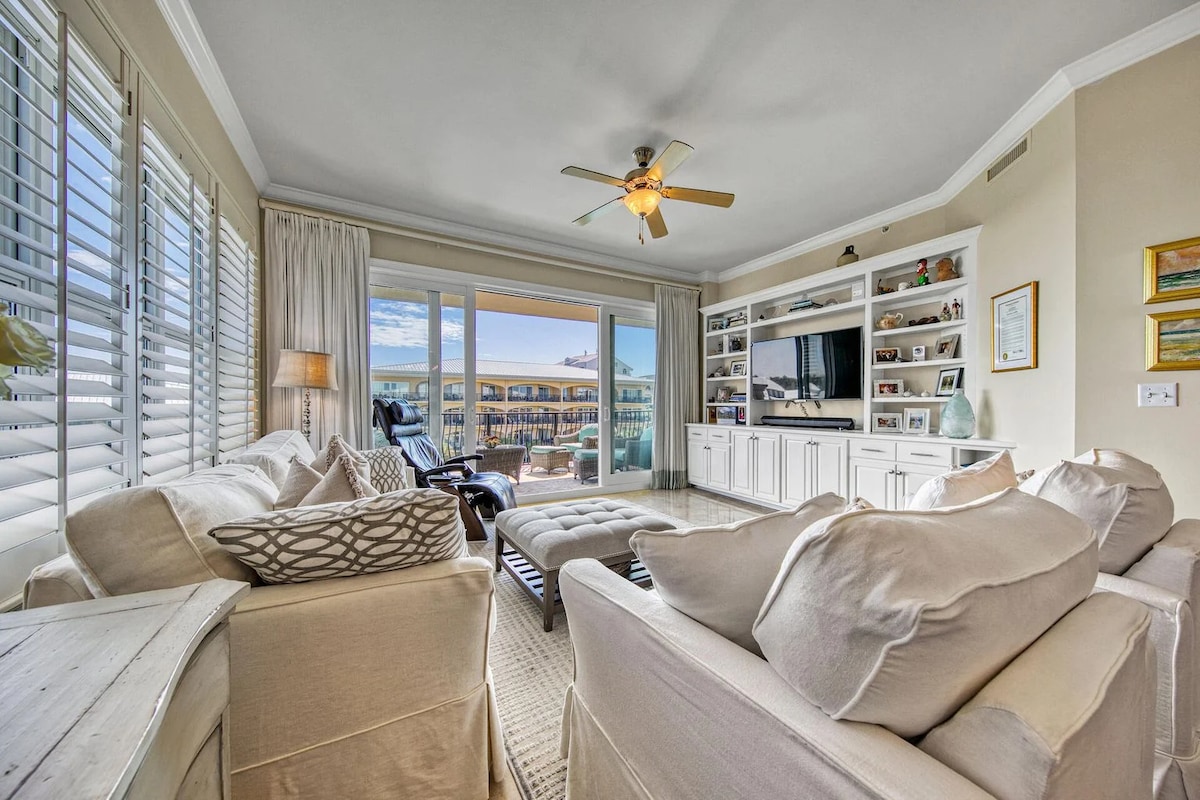 A spacious living area is showcased, featuring comfortable seating arranged around a coffee table. Large glass doors lead to scenic views outside, while built-in shelving adds storage and display space. Soft natural light fills the room, complemented by a ceiling fan for ventilation.