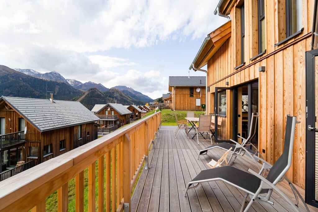 A wooden terrace is shown with sun loungers positioned for relaxation. The view opens to rolling hills and distant mountains, with wooden chalets in the background. Natural light enhances the warm tones of the wood, creating a welcoming outdoor space.
