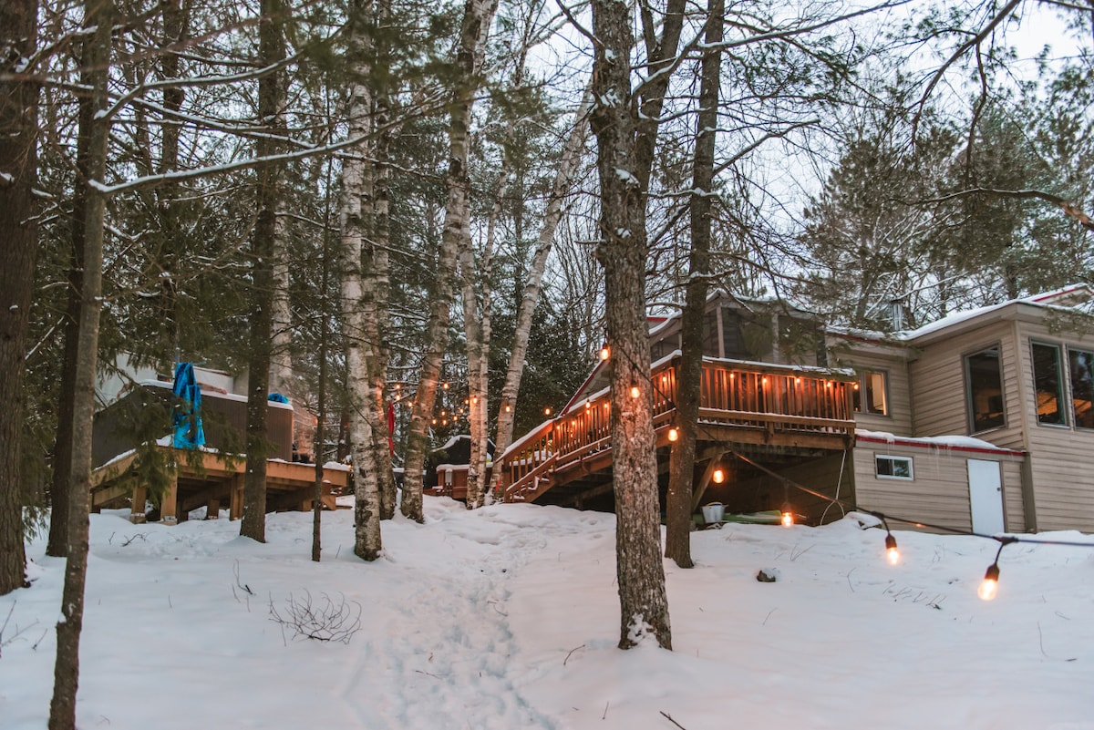 A charming cottage is nestled among snow-covered trees, featuring a gently sloping pathway leading to the entrance. Softly illuminated by string lights, the expansive deck invites relaxation, while the surrounding natural landscape captures the peaceful ambiance of the winter environment.