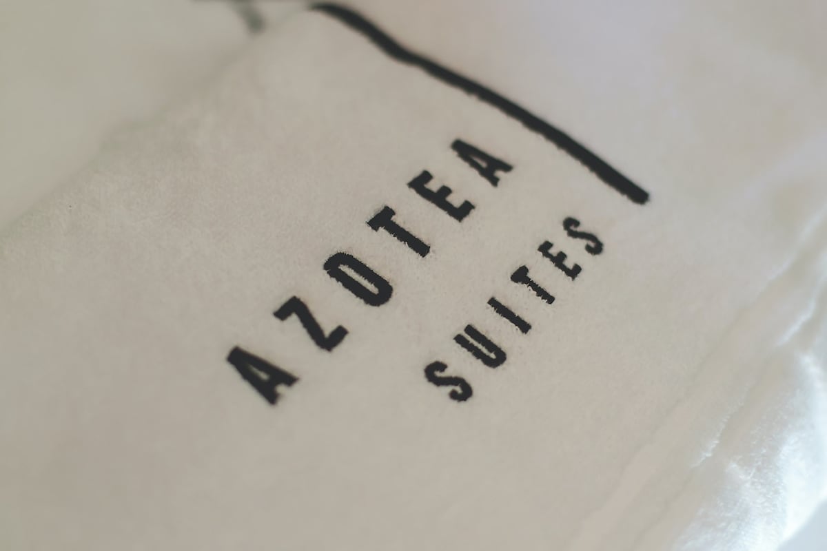 The image showcases a close-up of a plush towel with the elegant logo of Azotea Suites embroidered in black. The soft texture of the towel is highlighted, conveying a sense of luxury and comfort.