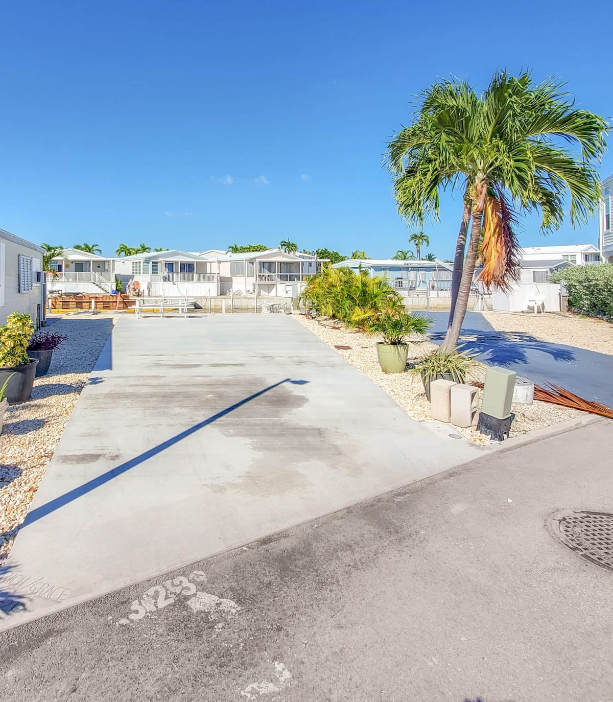 A concrete driveway leads toward RV lot access, framed by low-maintenance landscaping with stone and potted plants. Palm trees stand to the side, offering a touch of greenery against a clear blue sky. The area features well-maintained structures in the background.