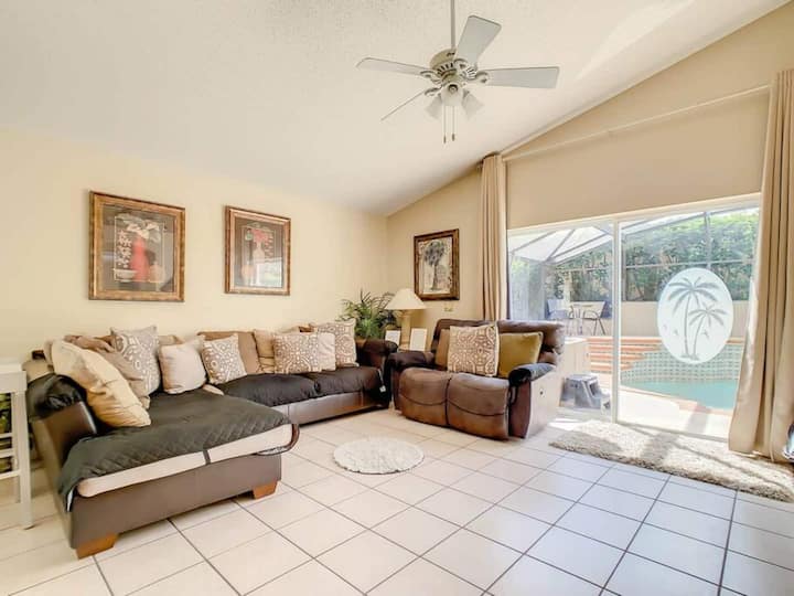 غرفة معيشة مع أريكة مقطعية وكرسي بظهر متحرك وباب زجاجي منزلق كبير يؤدي إلى منطقة حمام السباحة. الغرفة مزينة بألوان محايدة مع أعمال فنية على الجدران ومروحة سقف.