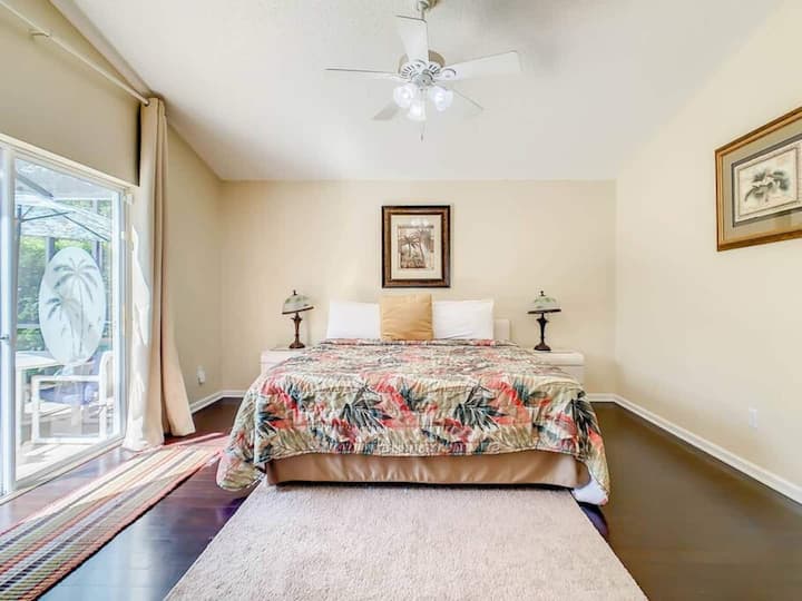 غرفة نوم رئيسية مع مروحة سقف، وباب زجاجي منزلق يؤدي إلى فناء. السرير مغطى بلحاف منقوش بالزهور، وهناك مصابيح على كلا الجانبين.