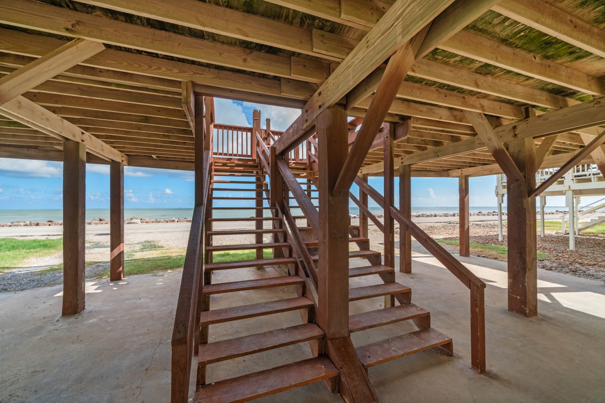 A wooden staircase leads to the upper level of the duplex, surrounded by a spacious area sheltered by the structure. The underside showcases wooden beams supporting the roof, with glimpses of beachfront visible in the background.
