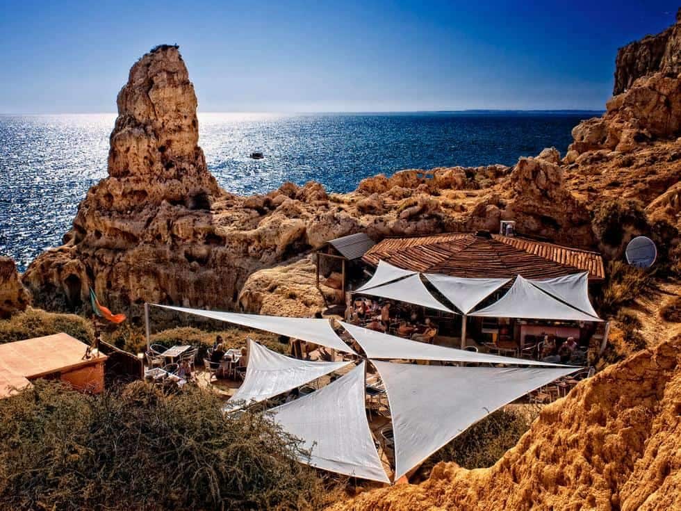 A coastal scene is captured featuring a rustic restaurant nestled among rocks, with ocean waves visible in the background. White canvas sails provide shade for outdoor seating areas, while the sun casts a warm glow over the landscape, highlighting the rugged terrain.
