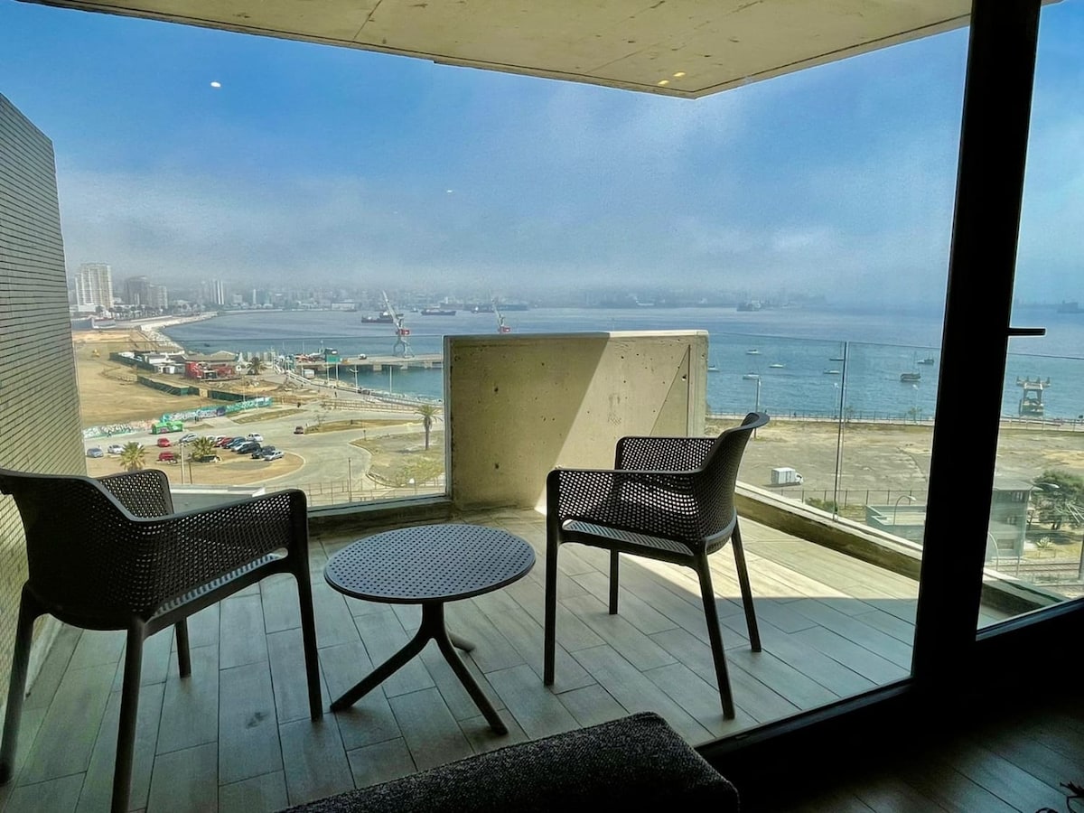 A private terrace features two modern chairs and a round table, offering a panoramic view of the city and the sea. The tranquil setting invites relaxation, with natural light flooding in through the large glass doors that connect the indoor space to the outdoor area.