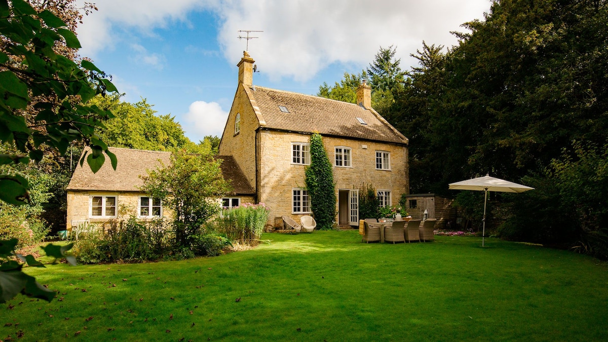 The cottage, constructed from mellow Cotswold stone, is set against a backdrop of lush greenery. It features a spacious garden with a manicured lawn, patio furniture, and a parasol, inviting the enjoyment of outdoor relaxation or gatherings.