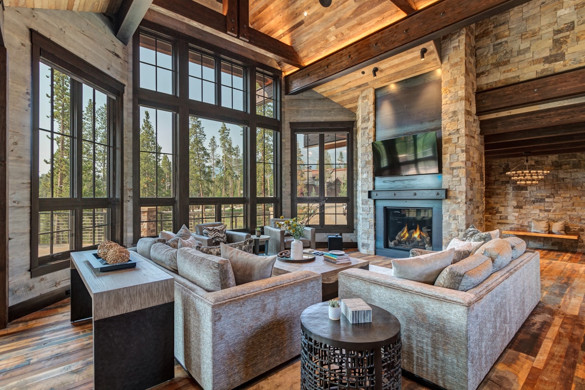 A spacious great room features large, tall windows framing views of surrounding evergreen trees. Cozy seating arrangements include multiple sofas and tables, complemented by a central stone fireplace. Warm wood elements are visible throughout, enhancing a comfortable setting.