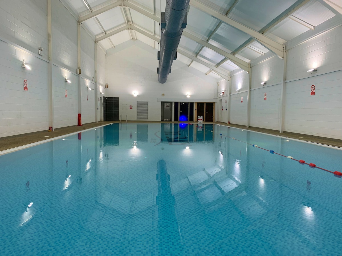 An indoor swimming pool is featured, showcasing clear blue water and tiled flooring. High ceilings and soft lighting create a spacious atmosphere, while safety markings are visible along the pool's edge. Entrance doors and changing facilities are located on the far wall.