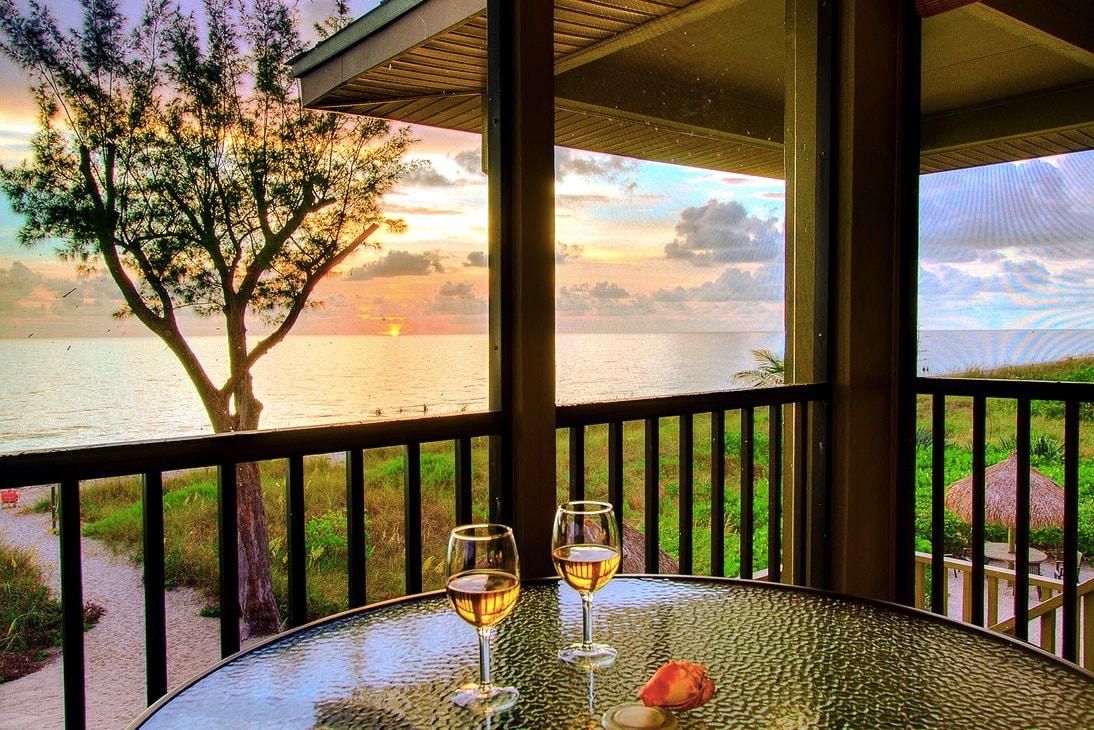 A glass table is set on a private lanai with two glasses of wine placed atop it. A serene view of the Gulf is captured beyond the railing, showcasing a sunset. Soft hues of orange and pink fill the sky, with a tree gently silhouetted in the foreground.
