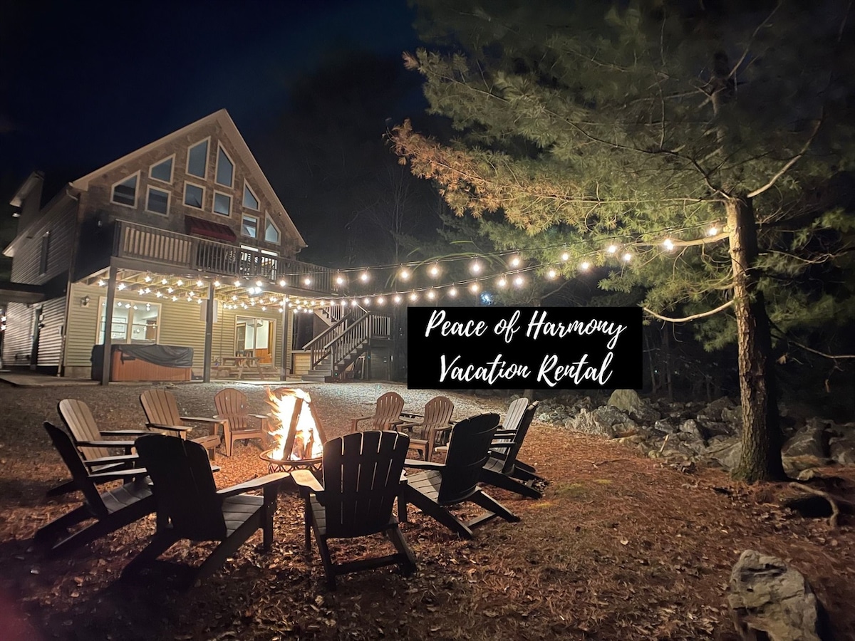 An outdoor gathering space is illuminated by string lights above. A fire pit with Adirondack chairs is surrounded by natural landscape, while the chalet's large windows reflect the warm glow. The rustic structure showcases traditional architectural features against a night backdrop.