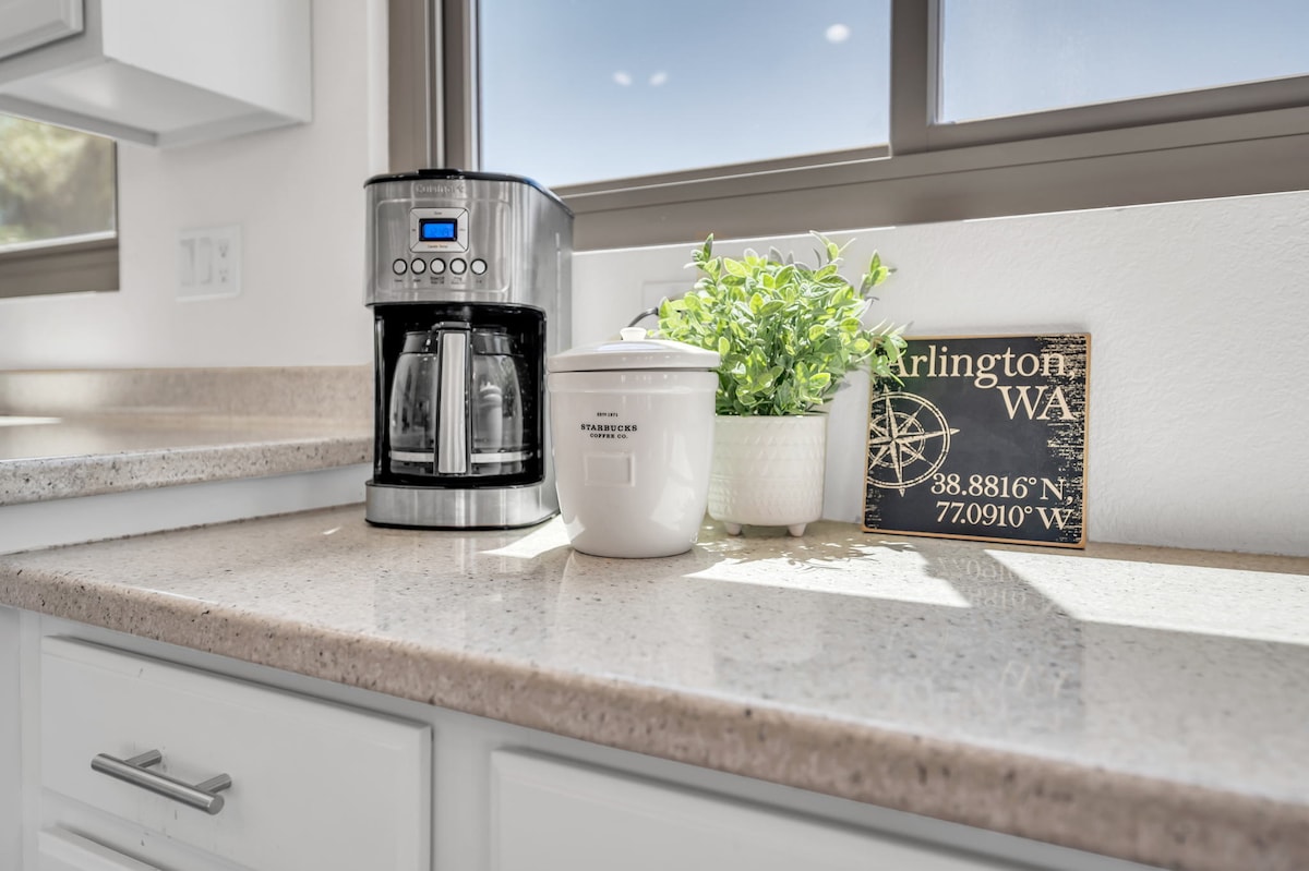 A modern kitchen countertop is shown, featuring a stainless-steel coffee maker with a digital display, a white ceramic container, and a small potted plant. Natural light filters through the nearby window, highlighting the sleek, light-colored countertop.
