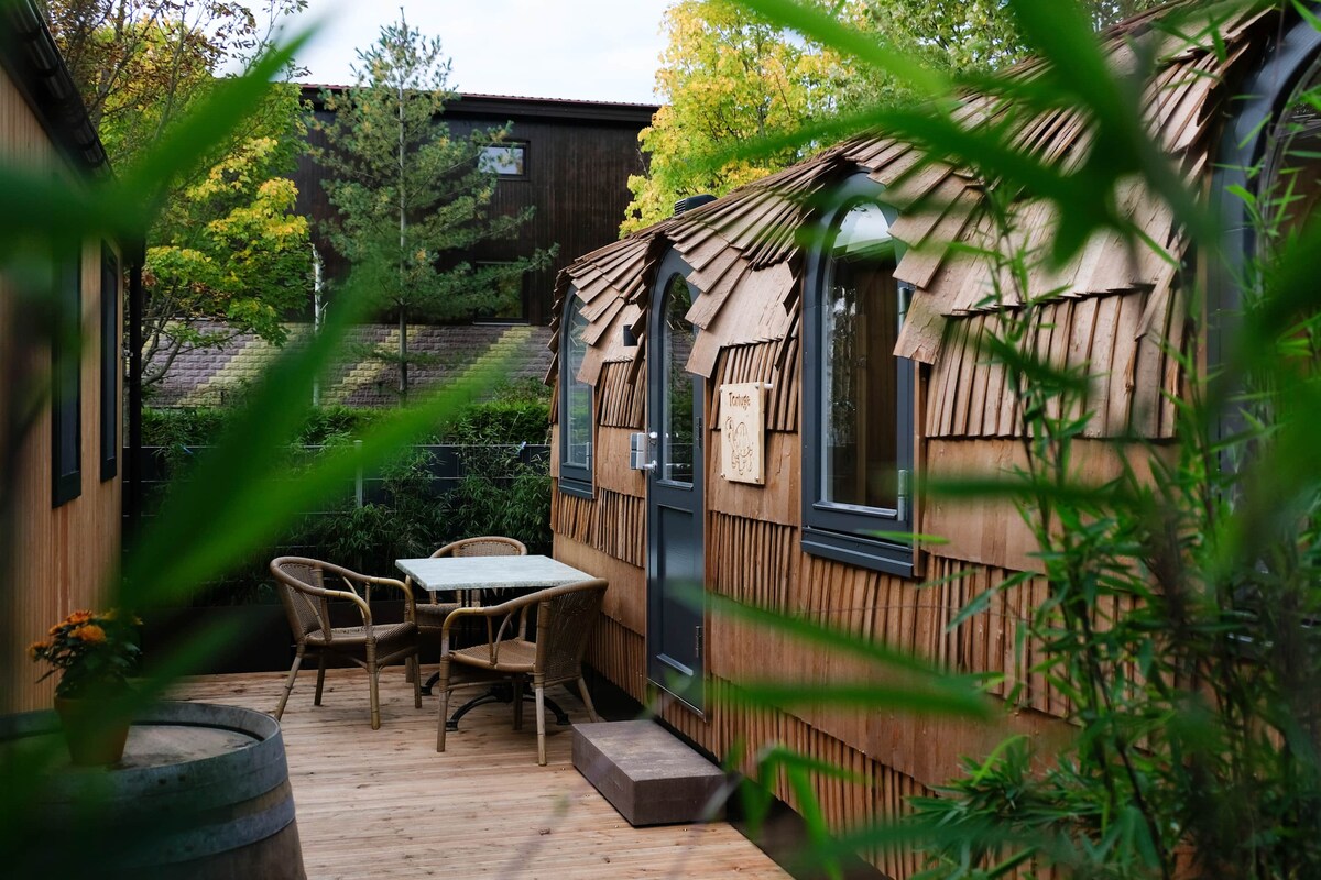 A quaint outdoor seating area is framed by lush greenery, featuring a wooden deck with a small table and chairs. The unique architecture of the nearby huts showcases a blend of wood shingles and dark window frames, set against a backdrop of trees.