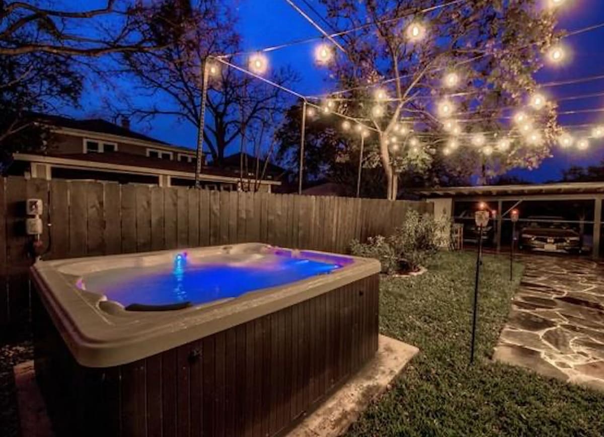 A hot tub is nestled in a fenced backyard, illuminated by string lights that create a relaxed ambiance. The inviting water glows with soft blue lighting. Nearby, a stone pathway and manicured grass offer a welcoming outdoor space.