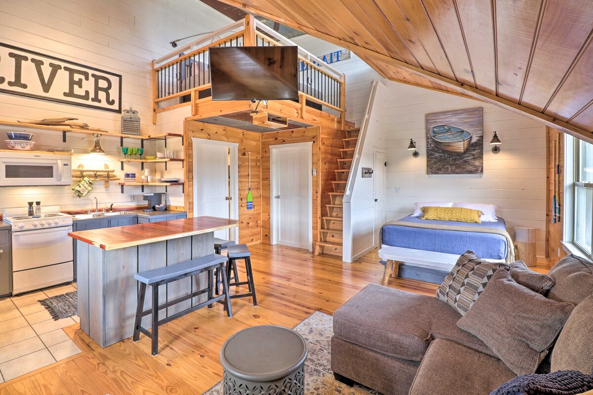 A spacious studio layout features a cozy seating area with a comfortable, textured couch. A sleek kitchen island with bar seating complements the wooden accents throughout the room. A lofted sleeping area is visible above, housing a queen bed with light linens.