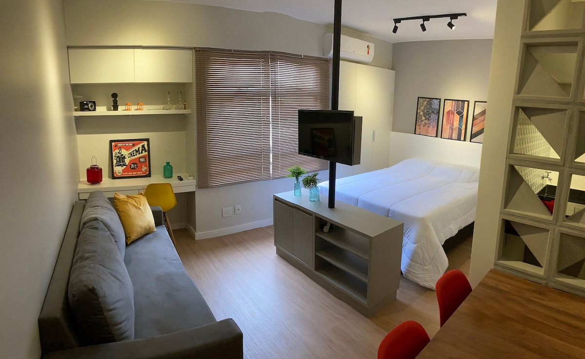 A modern studio space features a spacious bed with light linens, complemented by large windows with wooden shades. A grey sofa is positioned near a wooden dining table, while a mounted TV is visible on a central divider. Framed artwork adds a touch of character.