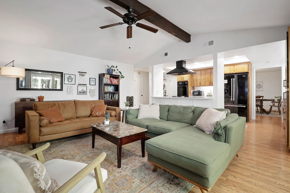 A spacious living area features a combination of comfortable seating, including a large green sectional and a beige sofa. Soft lighting illuminates the room, highlighting wooden beams and a decorative area rug. An open concept leads to a fully equipped kitchen in the background.