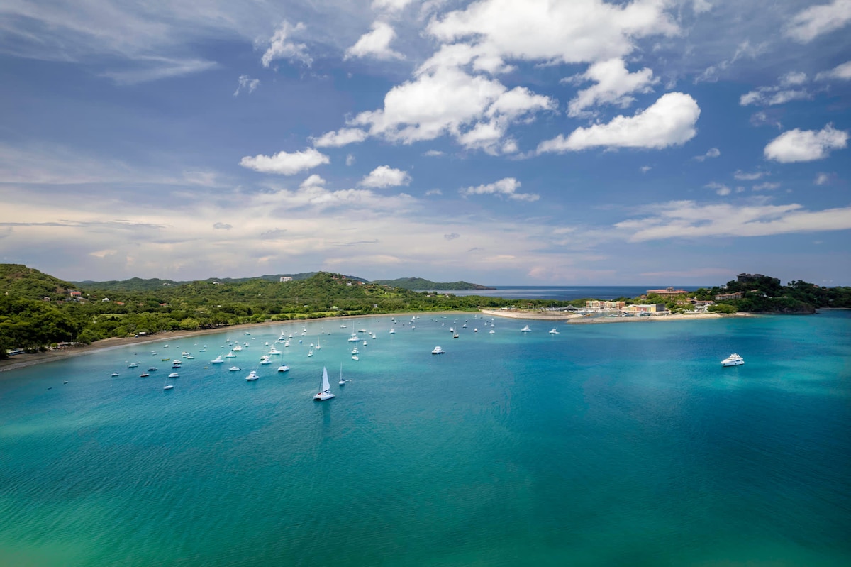 The image presents a stunning coastal view, featuring calm turquoise waters dotted with white sailboats. Lush greenery lines the shore, leading to distant hills under a partly cloudy sky. The serene landscape invites relaxation and exploration of nearby beaches and water activities.