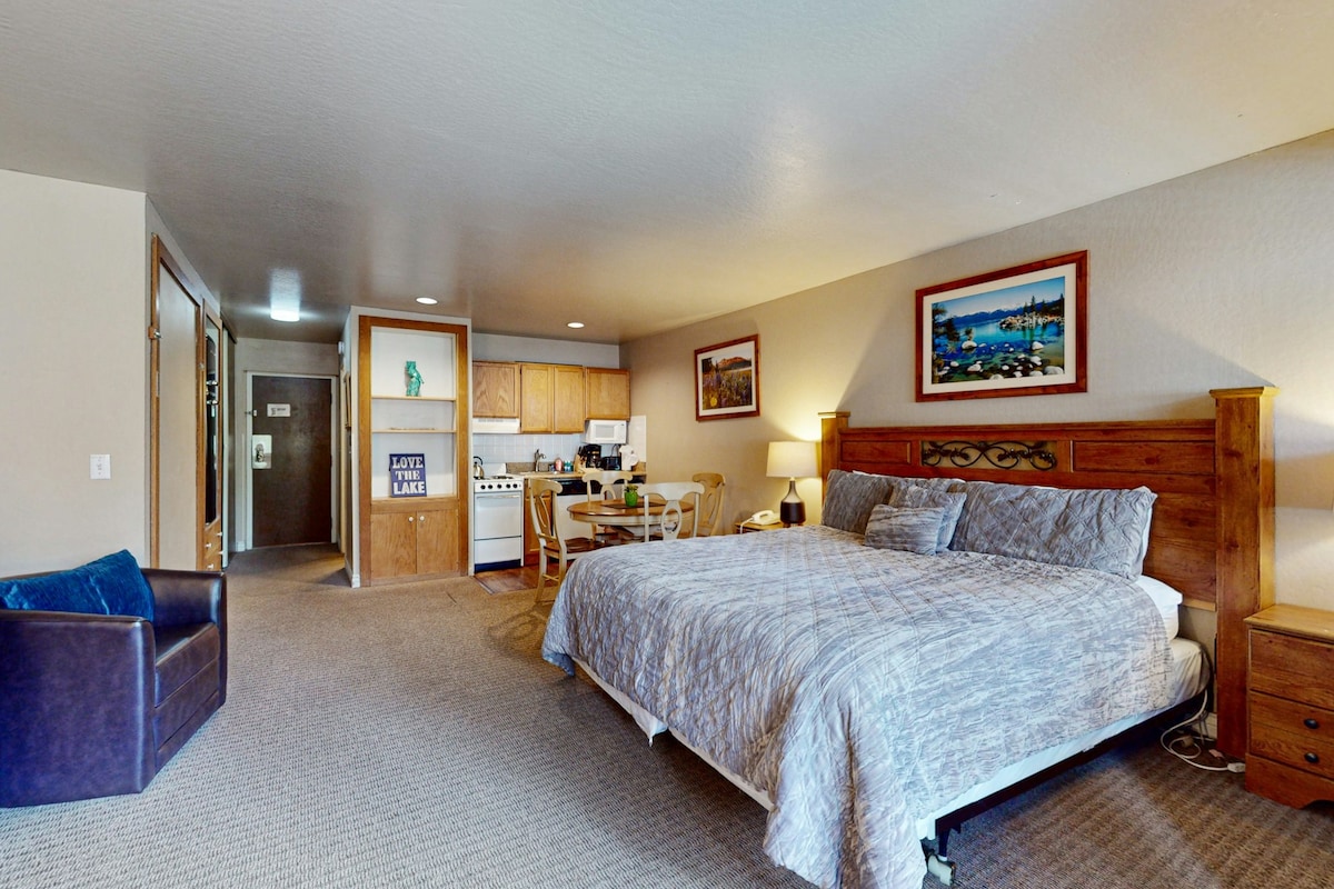 A spacious studio is presented with a large bed and a cozy chair. The kitchenette is visible in the background, equipped with essential appliances. Dining arrangements are provided at a small table with chairs. Bright artwork adorns the walls, contributing to the room's welcoming ambiance.