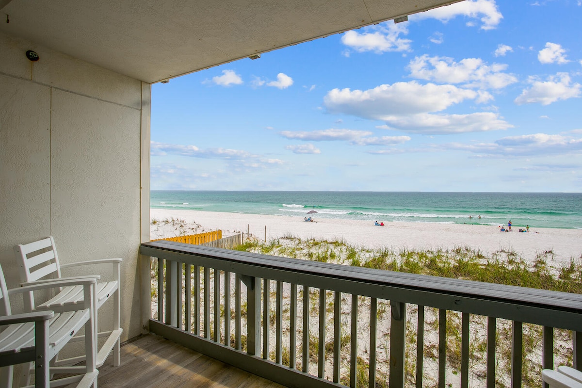 A private covered balcony overlooks the beach, featuring two white chairs positioned for ocean views. The foreground shows soft white sands leading to the emerald waters of the Gulf of Mexico, with a clear sky scattered with clouds in the background.