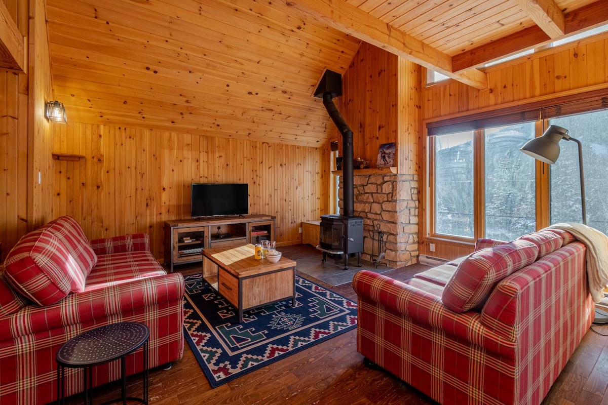 The living room features wooden paneling and high ceilings, creating a warm ambiance. Two plaid sofas are positioned around a coffee table, with a wood stove visible in the corner. Large windows provide natural light and views of the surrounding landscape.